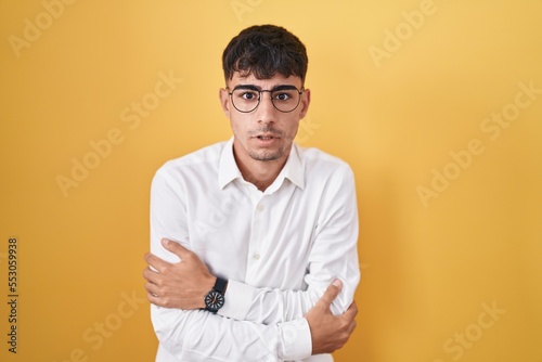 Young hispanic man standing over yellow background shaking and freezing for winter cold with sad and shock expression on face