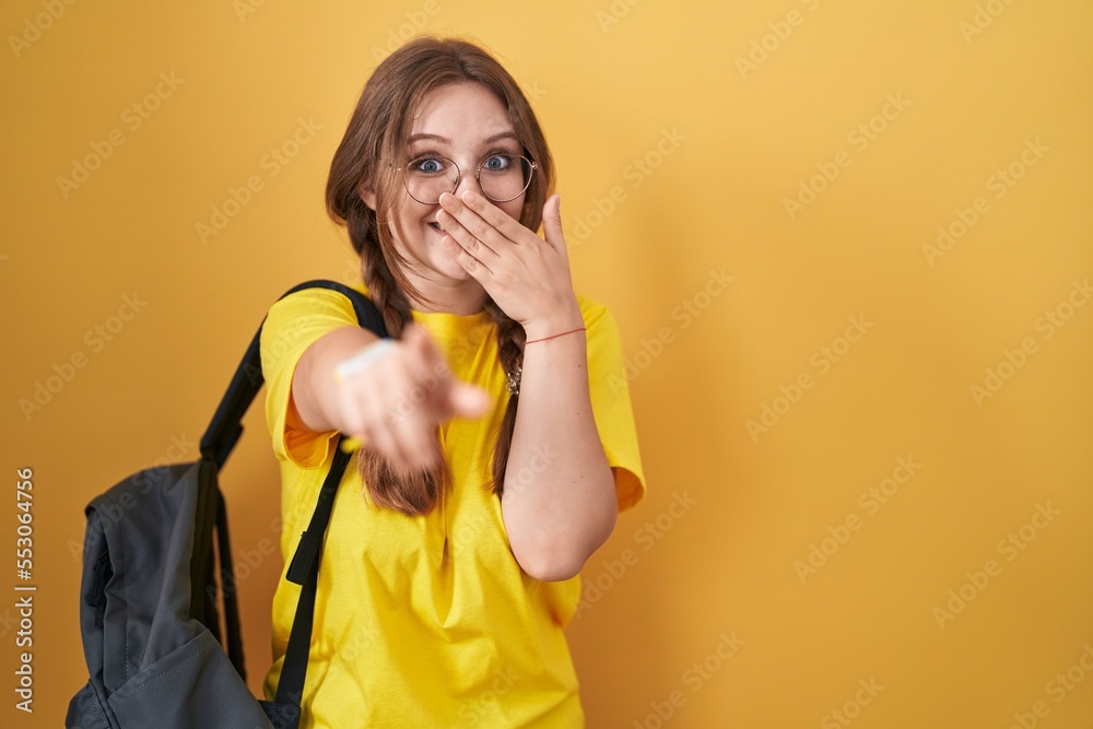 Young caucasian woman wearing student backpack over yellow background laughing at you, pointing finger to the camera with hand over mouth, shame expression