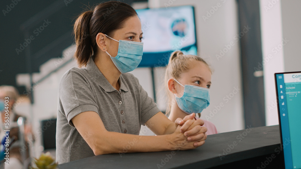 Mother and child with face mask talking to receptionist at hospital counter, people having medical appointment to see general practitioner. Woman and girl waiting at reception desk. Handheld shot.