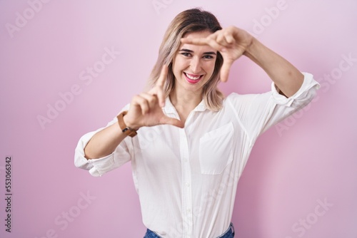 Young beautiful woman standing over pink background smiling making frame with hands and fingers with happy face. creativity and photography concept.