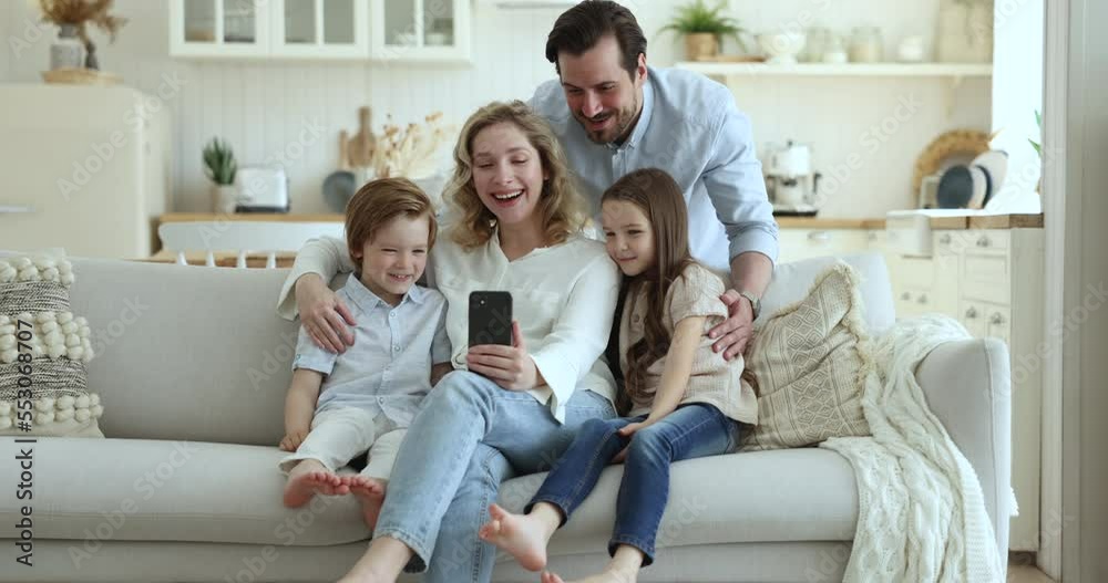 Happy attractive couple with little kids having fun use smartphone sit on cozy sofa in living room looking at device screen enjoy new mobile amusing application. Leisure of family using modern tech Happy attractive couple with little kids having fun use smartphone sit on cozy sofa in living room looking at device screen enjoy new mobile amusing application. Leisure of family using modern tech