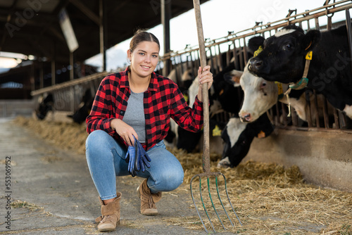 Portrait of positive young woman farmer with pitchfork squatting in cowshed among rows of cows in stalls.