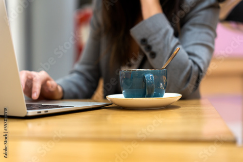 Crop anonymous female entrepreneur with long dark hair in gray suit working on laptop while sitting at wooden table with cup of cappuccino