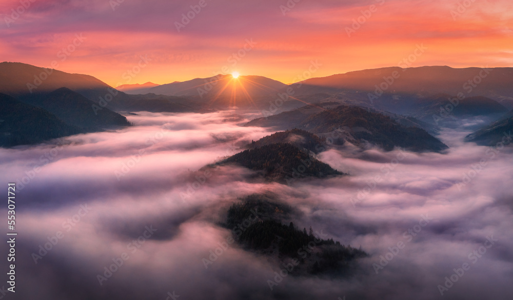 Aerial view of mountains in low clouds at sunrise in autumn. Top view of mountain hills in fog in fall at dawn. Beautiful landscape with ridges, forest, sun, colorful orange sky with pink clouds