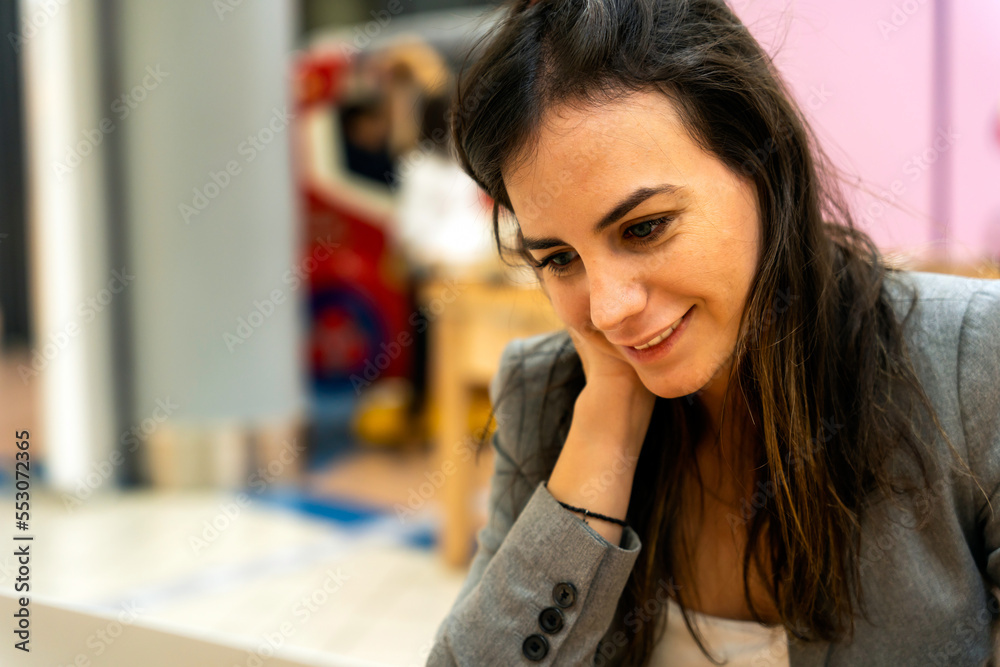 Positive adult female employee with long dark hair in formal suit smiling while sitting at table with hand on cheek Positive adult female employee with long dark hair in formal suit smiling while sitting at table with hand on cheek