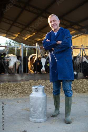 Positive middle-aged man farmer standing in cowshed with arms crossed at milk can and smiling.