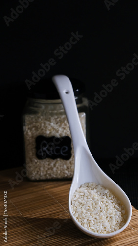 Tarro con granos de arroz y cucharón blanco con arroz en fondo negro. Dark food photo