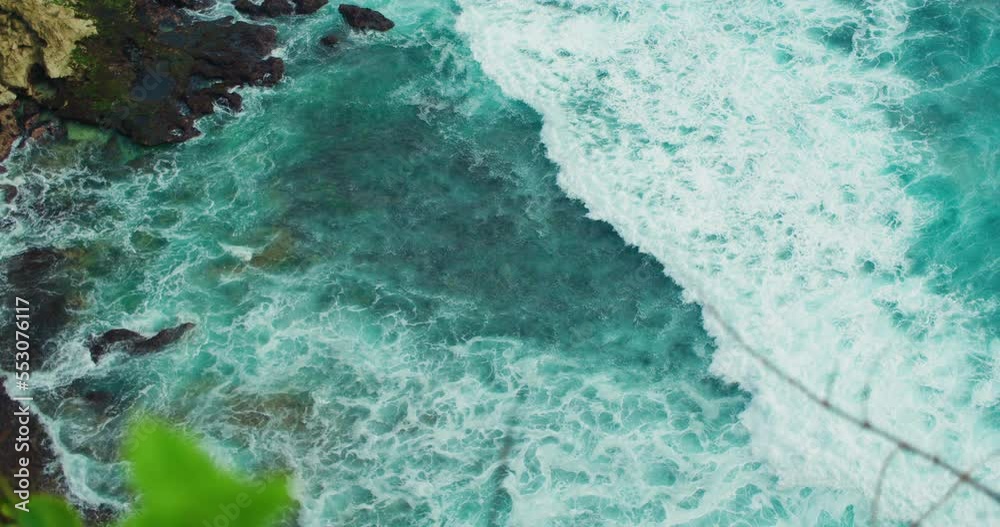 Top down static view from above of giant ocean waves crashing and foaming on empty rocky tropical beach with big cliff limestone stones. Wild shoreline Uluwatu Bali meeting deep blue sea water. Top down static view from above of giant ocean waves crashing and foaming on empty rocky tropical beach with big cliff limestone stones. Wild shoreline Uluwatu Bali meeting deep blue sea water.