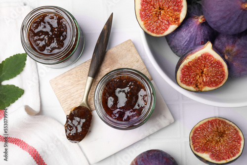 Glass jars of tasty sweet fig jam and fruits on white tiled table, flat lay © New Africa