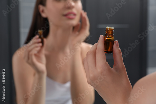 Young woman applying essential oil onto face indoors, closeup