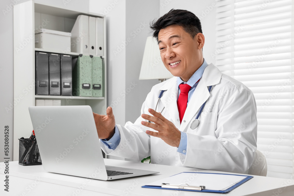 Obraz premium Happy doctor with laptop consulting patient at white desk in clinic. Online medicine