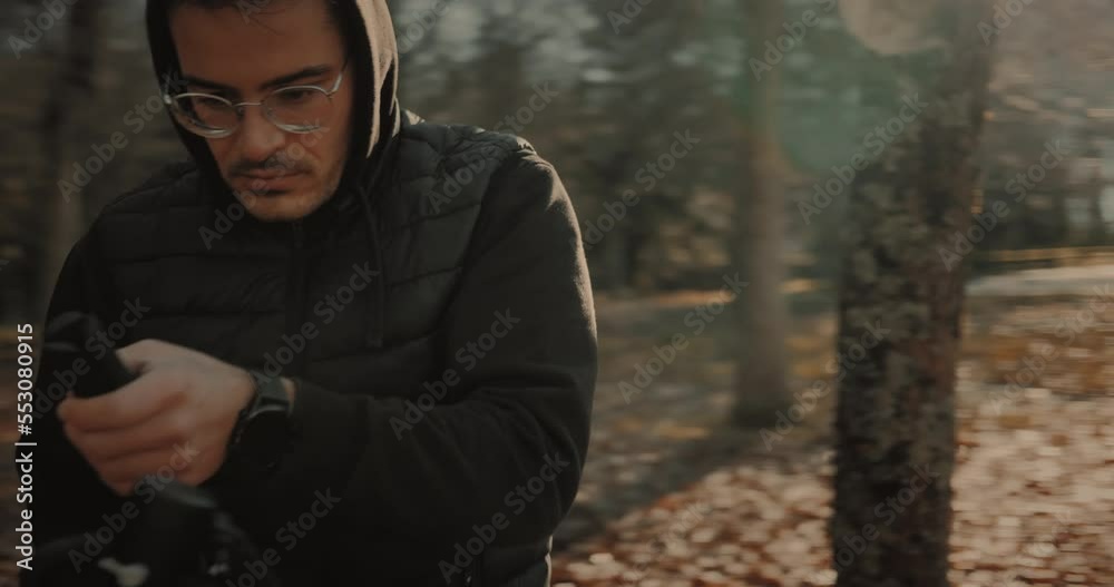 Focused young Caucasian male videomaker wearing glasses, black vest, and hoodie, working outdoors and recording video at sunset using a DSLR mirrorless camera mounted on a motorized stabilizer gimbal.