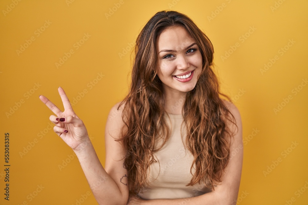 Young hispanic woman standing over yellow background smiling with happy face winking at the camera doing victory sign. number two. Young hispanic woman standing over yellow background smiling with happy face winking at the camera doing victory sign. number two.