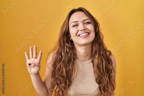 Young hispanic woman standing over yellow background showing and pointing up with fingers number four while smiling confident and happy.