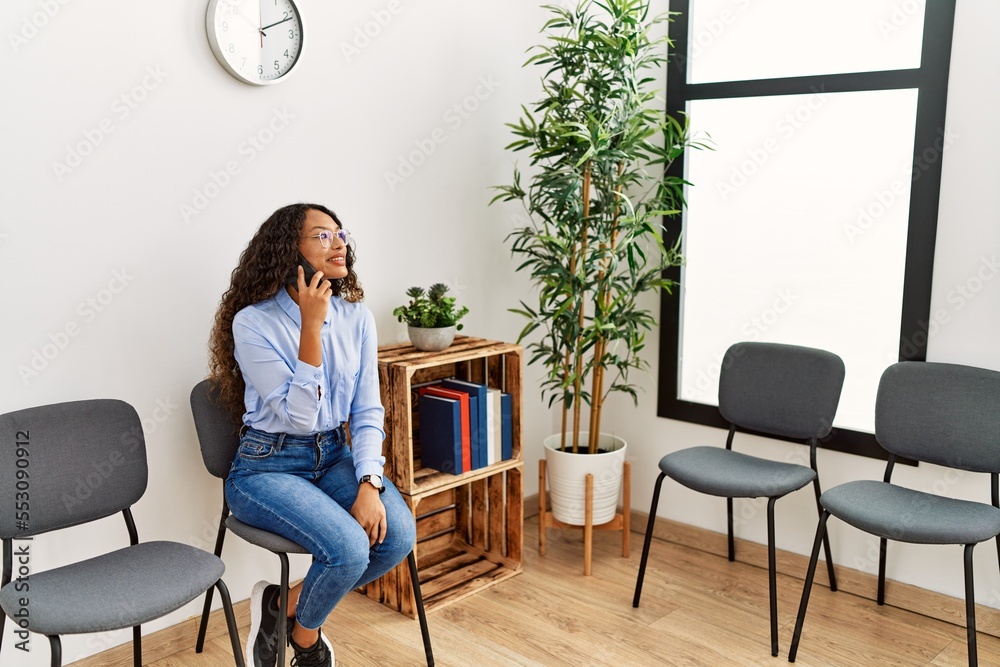 Young latin woman talking on the smartphone sitting on chair at waiting room
