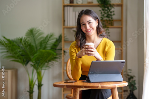 Pretty Asian woman sitting happily working with laptop and drinking coffee smiling and laughing brightly on a comfortable day at home.