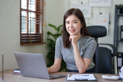 Attractive Asian businesswoman enjoying working using laptop at office. Looking at camera.