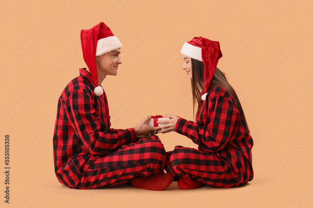 Φωτογραφία Young couple in Santa hats and pajamas with Christmas gift on beige background