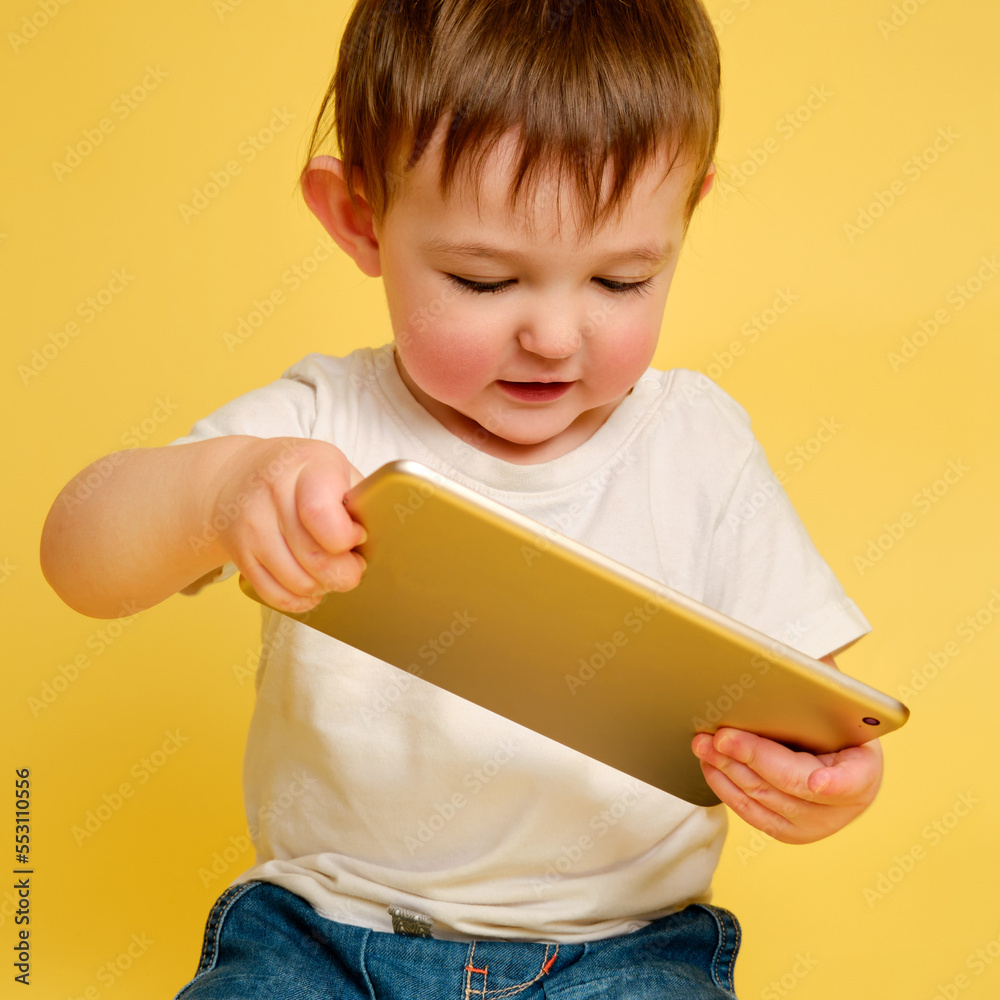 Toddler baby with a digital tablet on a studio yellow background. Happy child playing with a tablet in a white t-shirt and blue jeans. Kid aged one year and four months Toddler baby with a digital tablet on a studio yellow background. Happy child playing with a tablet in a white t-shirt and blue jeans. Kid aged one year and four months