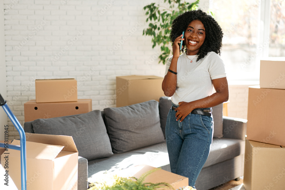 Young african woman with smartphone and boxes moving to new home