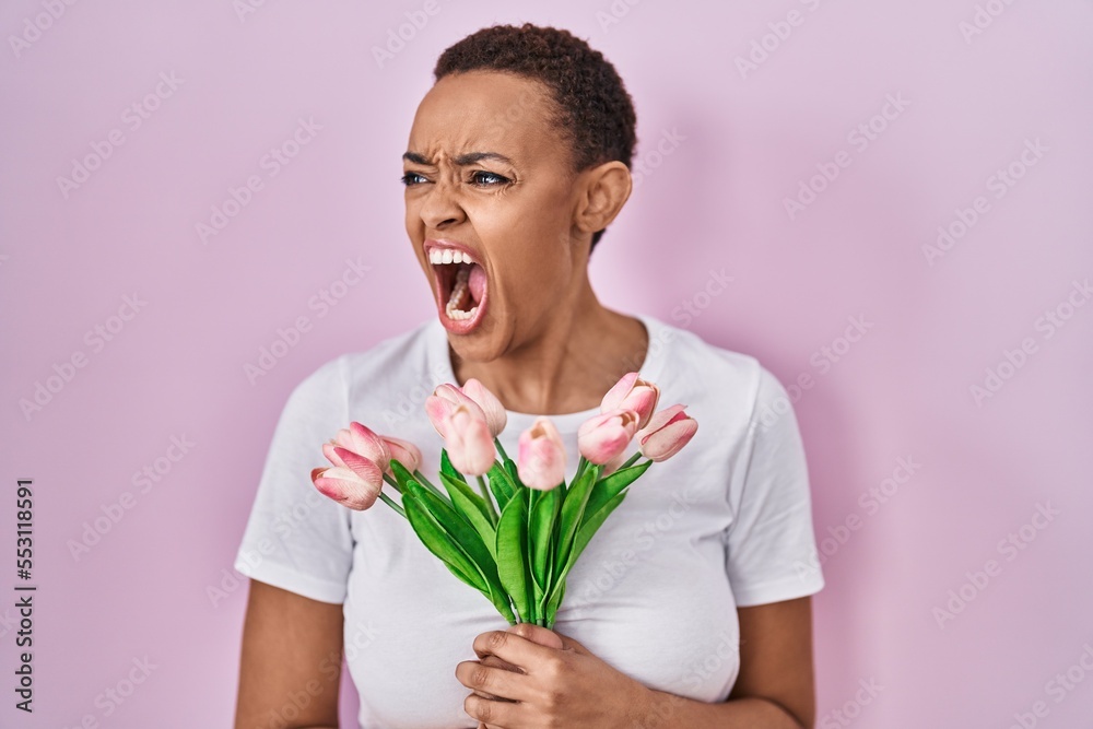 Beautiful african american woman holding bouquet of pink tulips flowers angry and mad screaming frustrated and furious, shouting with anger. rage and aggressive concept. Beautiful african american woman holding bouquet of pink tulips flowers angry and mad screaming frustrated and furious, shouting with anger. rage and aggressive concept.