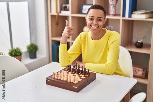 African american woman playing chess sitting on the table smiling happy and positive, thumb up doing excellent and approval sign