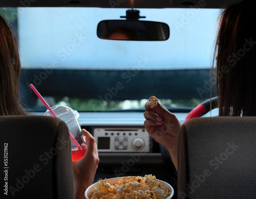 Two female friends with popcorn sitting in the car while watching a movie at drive in cinema. Selective focus. Entertainment, leisure activities, hobby concept