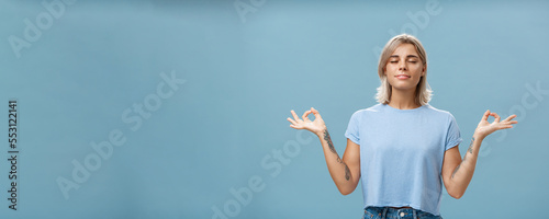 Listening to inner self during meditation. Calm happy and relaxed attractive tanned blonde female with tattoos on arms standing in lotus pose over blue wall with zen gesture closed eyes doing yoga