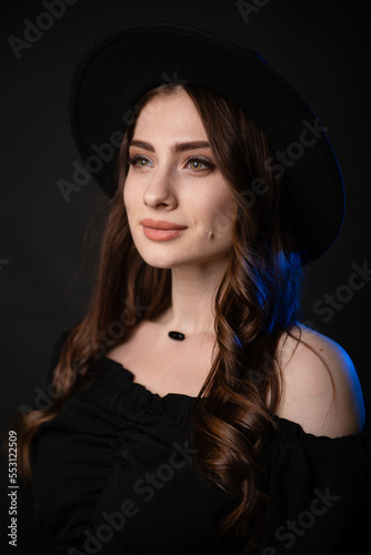 Beautiful young woman in black clothes with a black hat. Photo in the studio on a dark background with blue backlight.