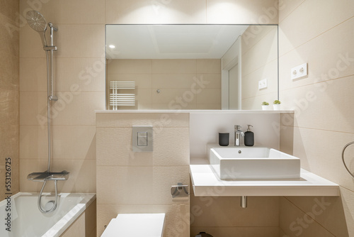 Cozy bathroom in modern design with huge mirror on the wall and bright lighting. Walls are in beige tiles and white sanitary ware. Room for taking morning shower and relaxing evening baths.