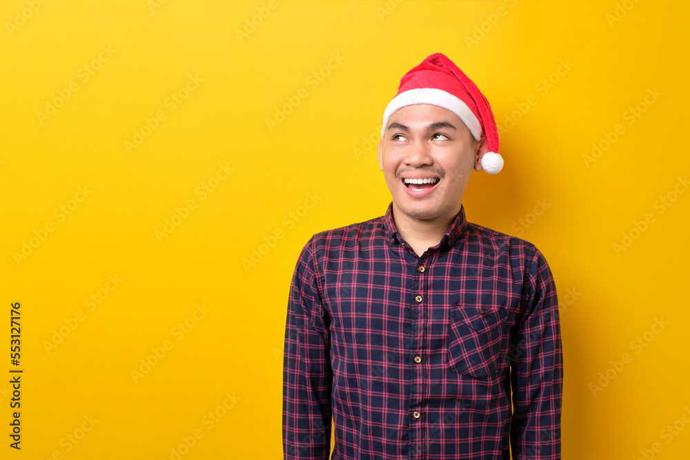 Happy young Asian man in Santa hat looking upwards with broad smile on yellow studio background. Happy New Year 2023 celebration merry holiday concept
