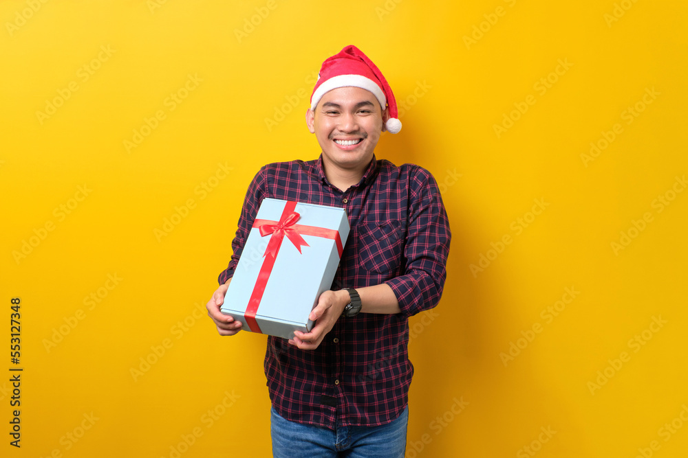 Cheerful young Asian man in Santa hat holding gift box with ribbon on yellow studio background. Happy New Year 2023 celebration merry holiday concept Cheerful young Asian man in Santa hat holding gift box with ribbon on yellow studio background. Happy New Year 2023 celebration merry holiday concept