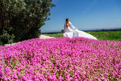 a beautiful woman in a white flowing long dress stay near a beautiful field with pink flowers rear view