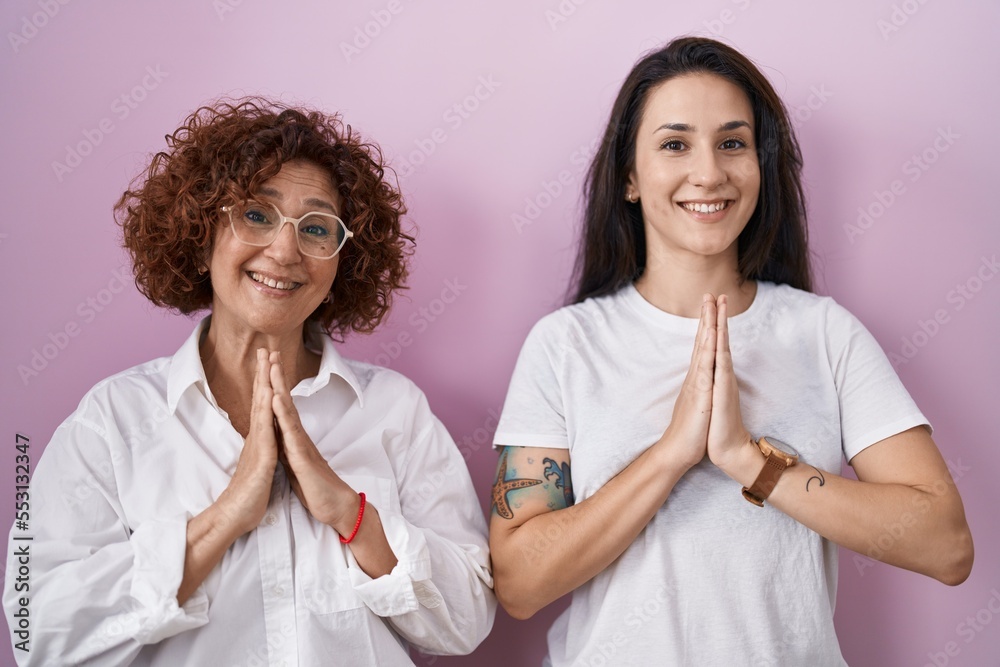Hispanic mother and daughter wearing casual white t shirt over pink background praying with hands together asking for forgiveness smiling confident.