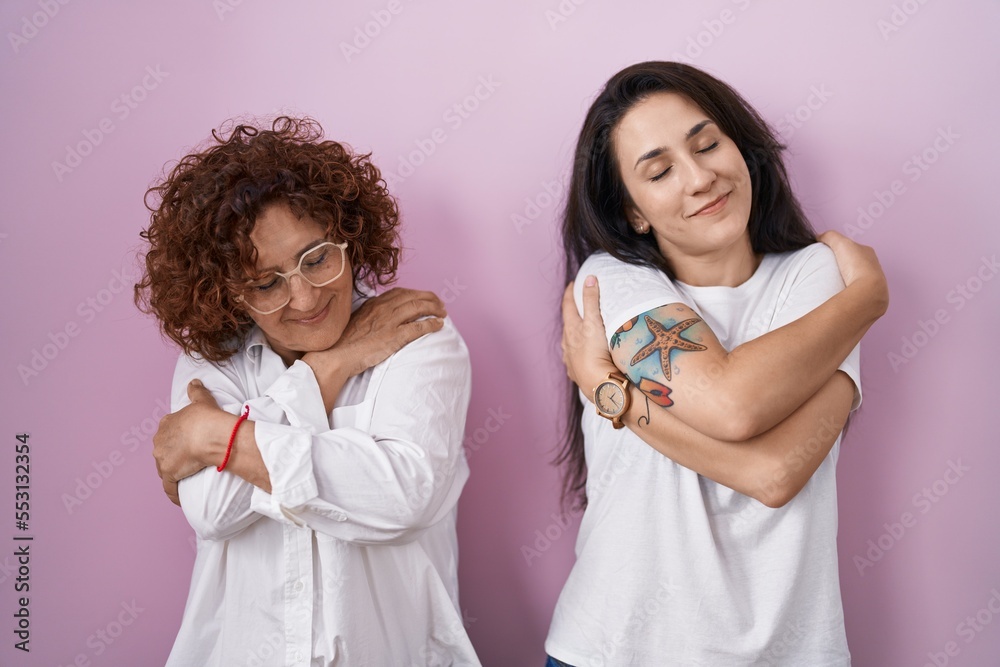 Hispanic mother and daughter wearing casual white t shirt over pink background hugging oneself happy and positive, smiling confident. self love and self care Hispanic mother and daughter wearing casual white t shirt over pink background hugging oneself happy and positive, smiling confident. self love and self care
