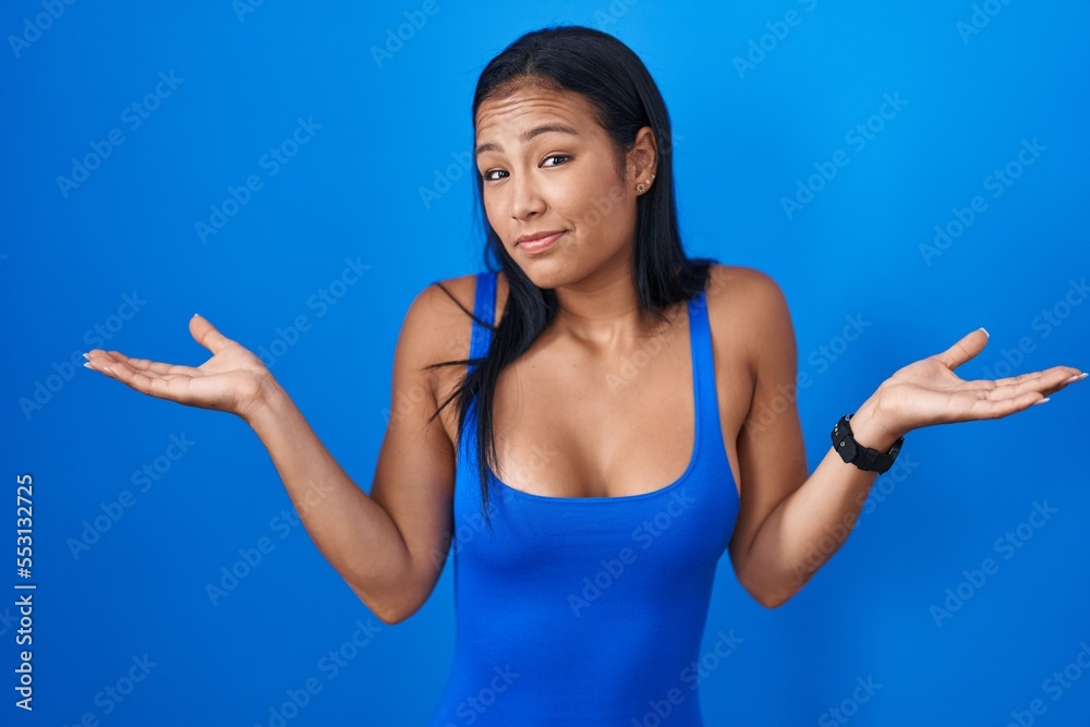 Hispanic woman standing over blue background clueless and confused expression with arms and hands raised. doubt concept. Hispanic woman standing over blue background clueless and confused expression with arms and hands raised. doubt concept.