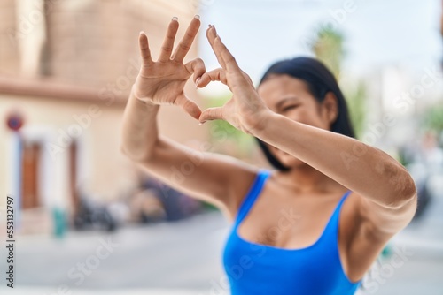 Young beautiful latin woman smiling confident doing heart gesture with hands at street