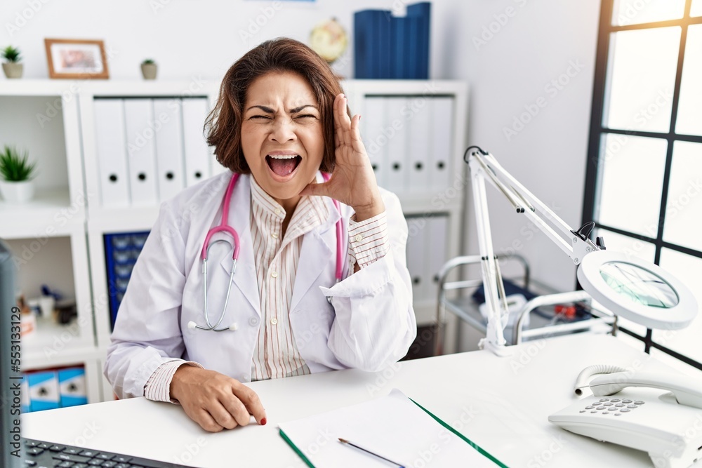 Middle age hispanic woman wearing doctor uniform and stethoscope at the clinic shouting and screaming loud to side with hand on mouth. communication concept. Middle age hispanic woman wearing doctor uniform and stethoscope at the clinic shouting and screaming loud to side with hand on mouth. communication concept.