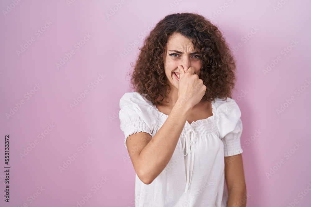 Hispanic woman with curly hair standing over pink background smelling something stinky and disgusting, intolerable smell, holding breath with fingers on nose. bad smell Hispanic woman with curly hair standing over pink background smelling something stinky and disgusting, intolerable smell, holding breath with fingers on nose. bad smell