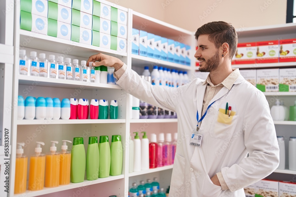 Young caucasian man pharmacist smiling confident holding product on shelving at pharmacy