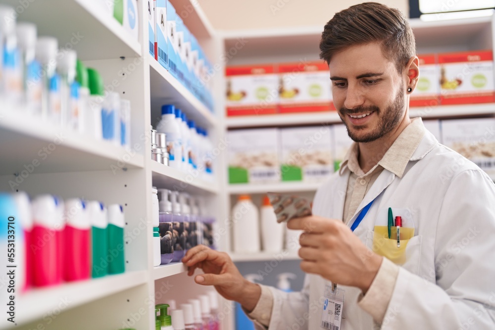 Young caucasian man pharmacist holding pills of shelving at pharmacy