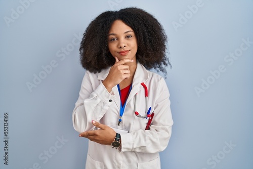 Young african american woman wearing doctor uniform and stethoscope looking confident at the camera smiling with crossed arms and hand raised on chin. thinking positive.