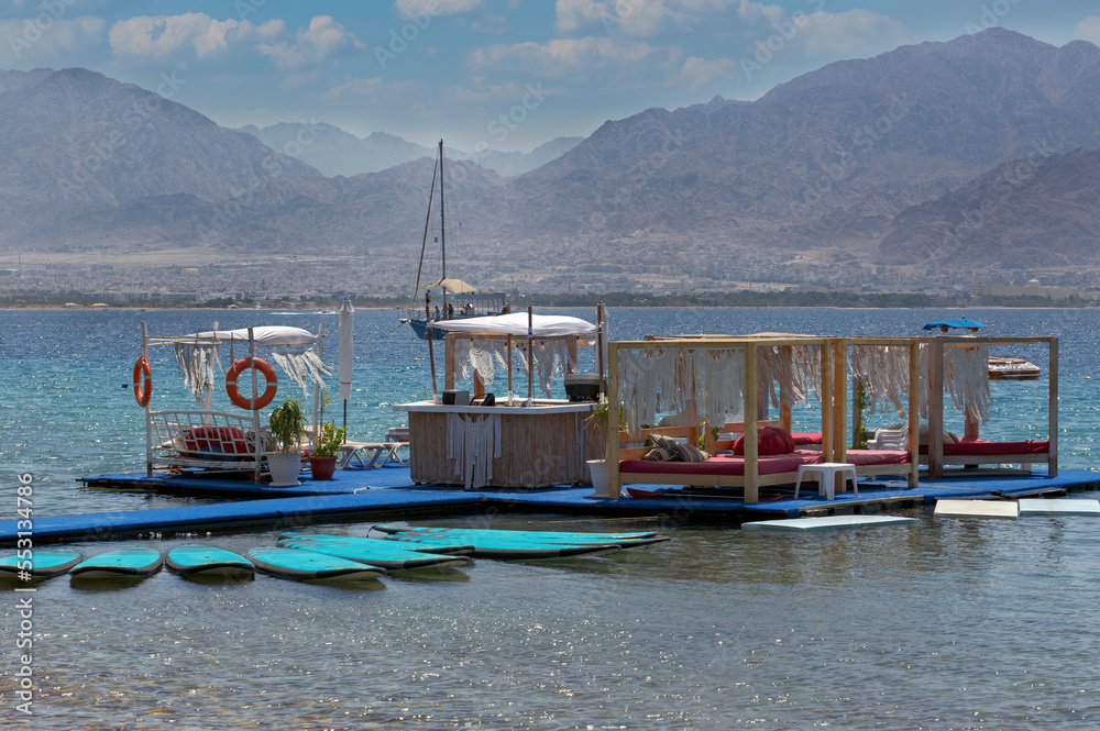 Traditional floating restaurants on the coast Israel. Empty floating restaurant and bar on Red sea at sunset. Surfboards on blue water. Travel adventure, water sport with sea and blue sky background