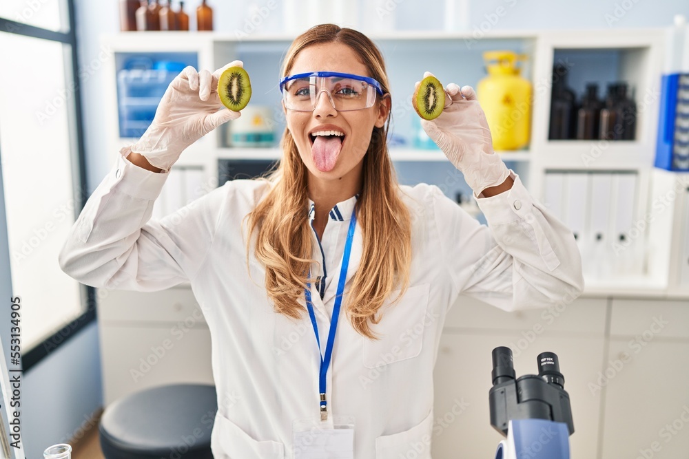 Young blonde scientist woman working with food at laboratory sticking tongue out happy with funny expression. Young blonde scientist woman working with food at laboratory sticking tongue out happy with funny expression.