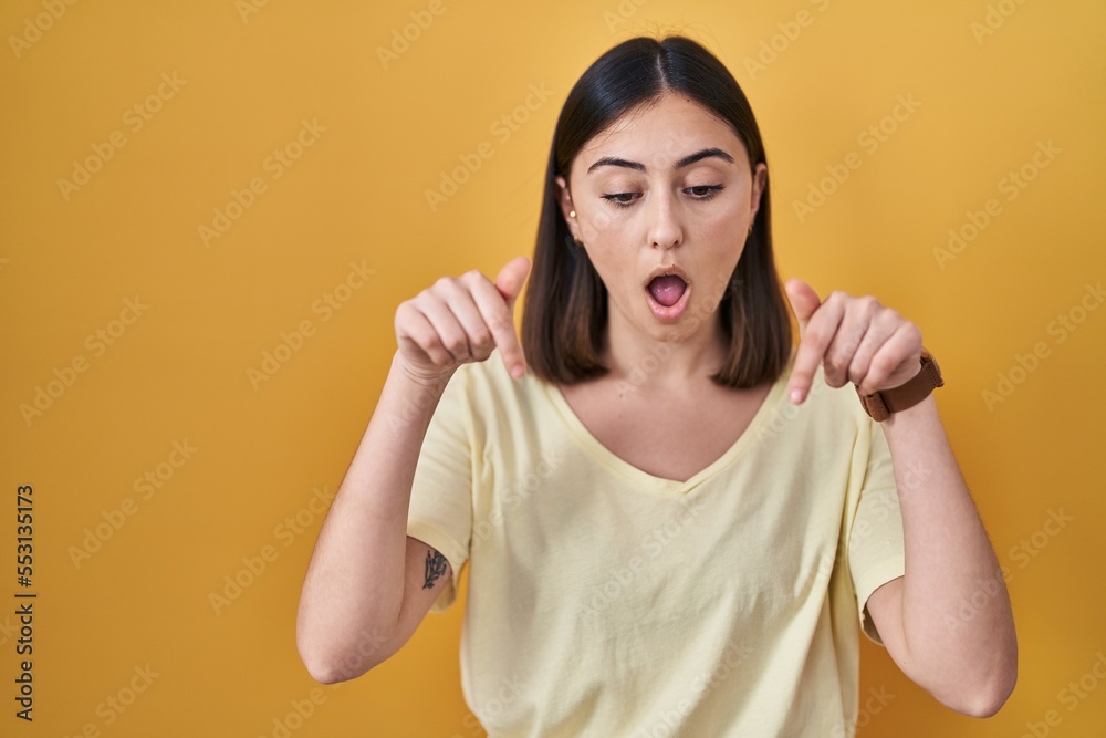 Hispanic girl wearing casual t shirt over yellow background pointing down with fingers showing advertisement, surprised face and open mouth Hispanic girl wearing casual t shirt over yellow background pointing down with fingers showing advertisement, surprised face and open mouth