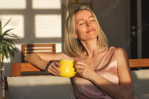 Happy dreamy middle aged woman sitting on sofa in living room with cup of tea or coffee photo