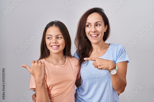 Young mother and daughter standing over white background smiling with happy face looking and pointing to the side with thumb up.
