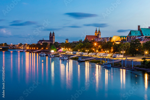 Germany, Saxony-Anhalt, Magdeburg, Boats on bank of Elbe river at dusk with city buildings in background photo