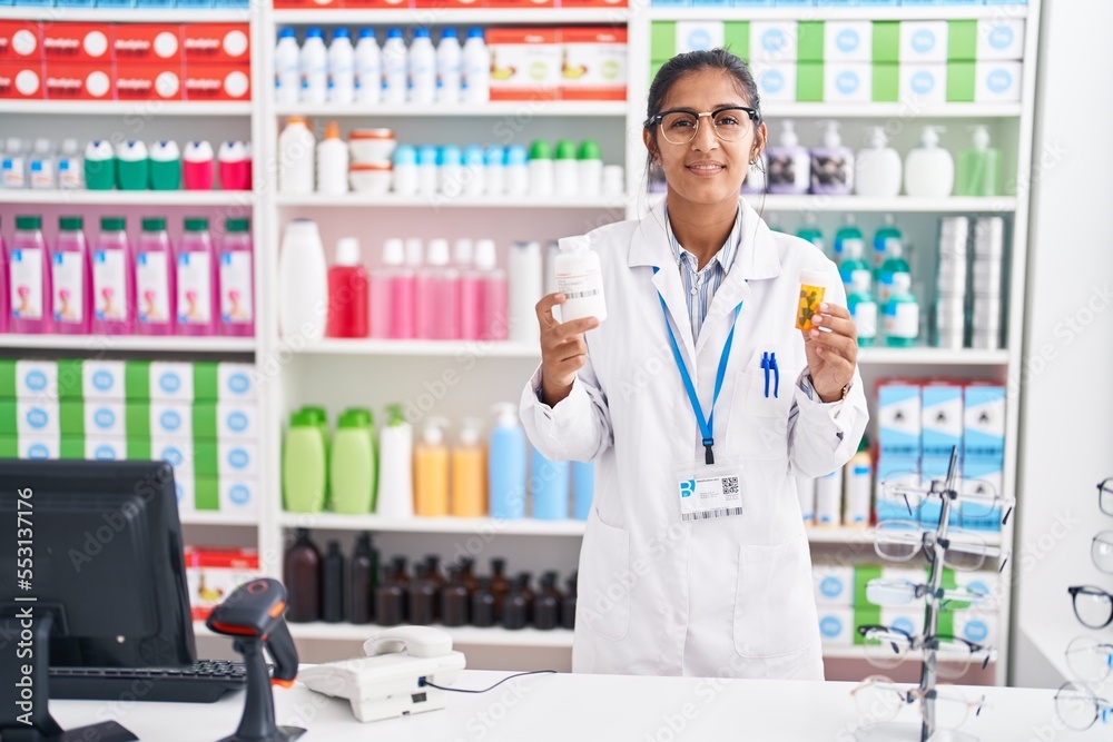 Young beautiful hispanic woman pharmacist smiling confident holding pills bottles at pharmacy