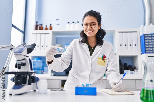 Young latin woman scientist writing on notebook holding pills at laboratory